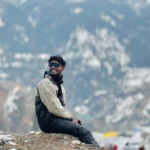 best travel photographer in wayanad A man sitting on a rocky snow-covered hillside, smiling and looking away, wearing winter clothing and reflective sunglasses, with snow-capped mountains and a blurred scenic valley in the background.